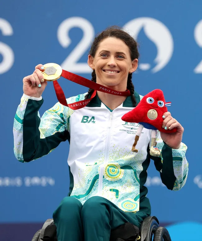 Australian Paralympian in wheelchair holding a gold medal and red plush mascot, smiling at the Paris 2024 event.