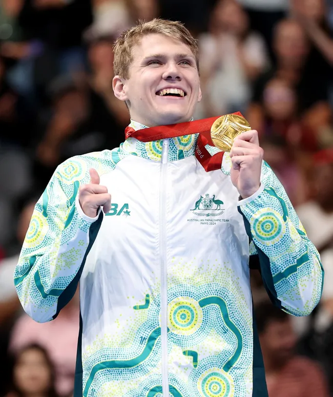 Australian Paralympic athlete smiling, holding a gold medal, wearing a patterned team jacket at a victory ceremony.