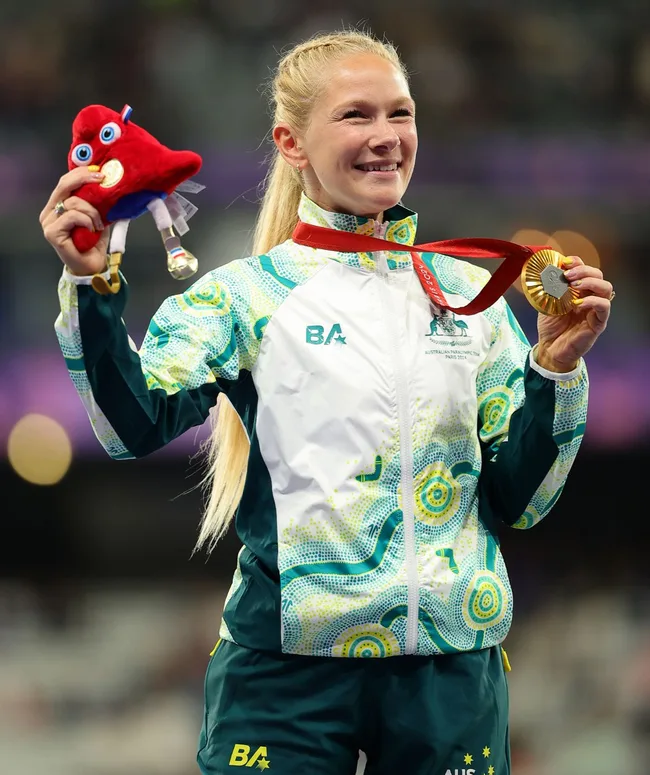 Australian athlete smiling, holding a toy and wearing a gold medal, in a patterned tracksuit with the initials "BA".