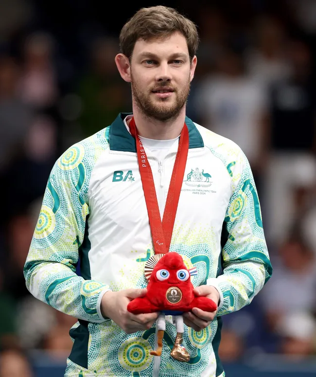 Person in an Australian Paralympic Team jacket holding a red mascot with a medal.