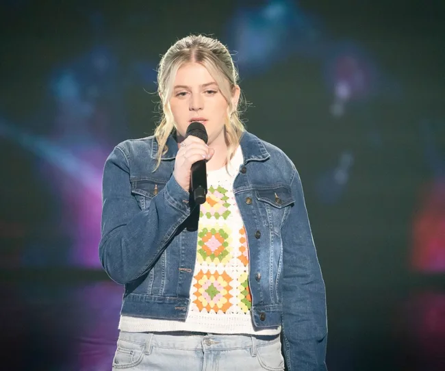 A person sings into a microphone, wearing a denim jacket and patterned sweater, against a blurred, colorful background.