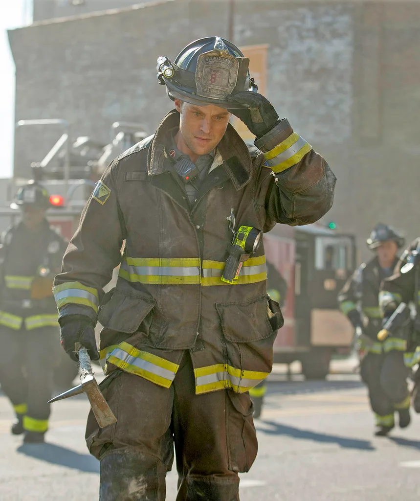 Jesse Spencer, dressed as a firefighter, walking down a smoky street with a firetruck behind him, in Chicago Fire.
