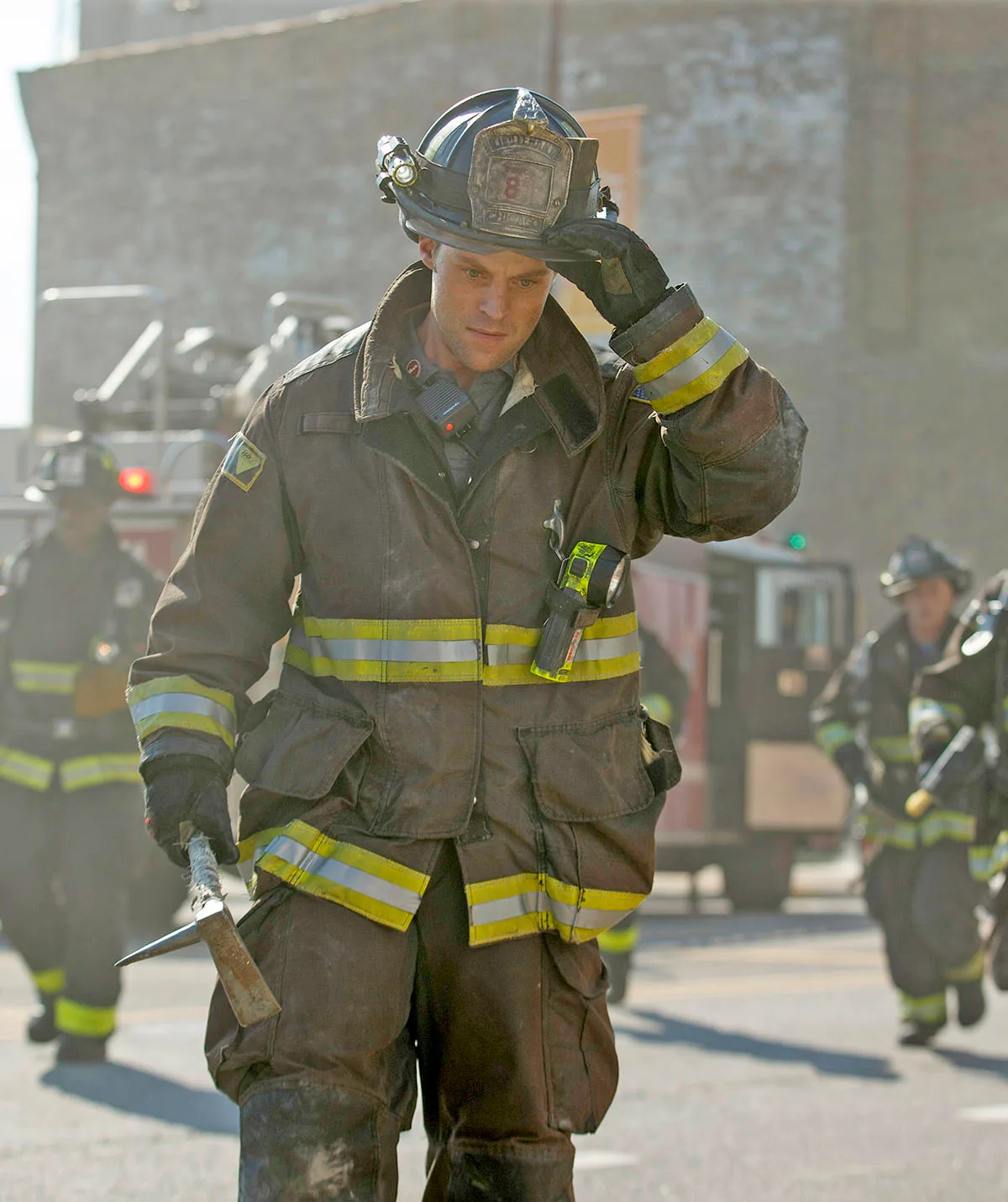 Jesse Spencer, dressed as a firefighter, walking down a smoky street with a firetruck behind him, in Chicago Fire.