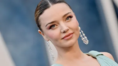 A woman with elegant earrings and a light blue dress smiles softly against a blurred background.