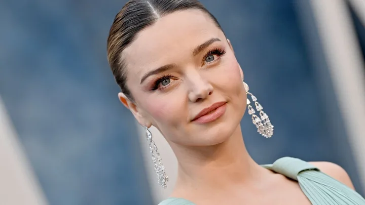 A woman with elegant earrings and a light blue dress smiles softly against a blurred background.