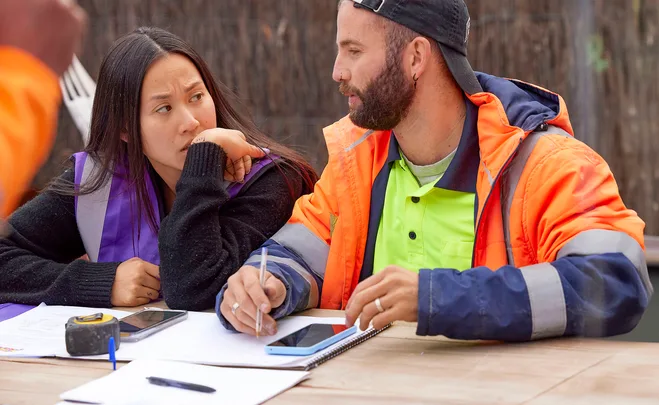 Mimi in purple hi vis and Kristian in orange hi vis sit at a table and look stressed over paperwork