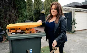 Terese hiding alcohol bottles in a full recycling bin on the side of the road while all dressed in black and looking suspicious