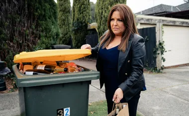 Terese hiding alcohol bottles in a full recycling bin on the side of the road while all dressed in black and looking suspicious