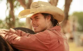 Sam Corlett in pink shirt and a cowboy hat leaning on a horse smiling