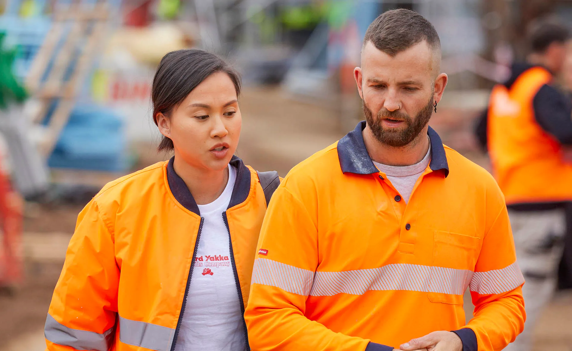 Mimi and Kristian walk in orange hi vis on site looking distressed