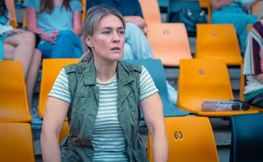 Jenni Baird, as Dana Hanlon in Plum, sits in a seat at a football stadium.