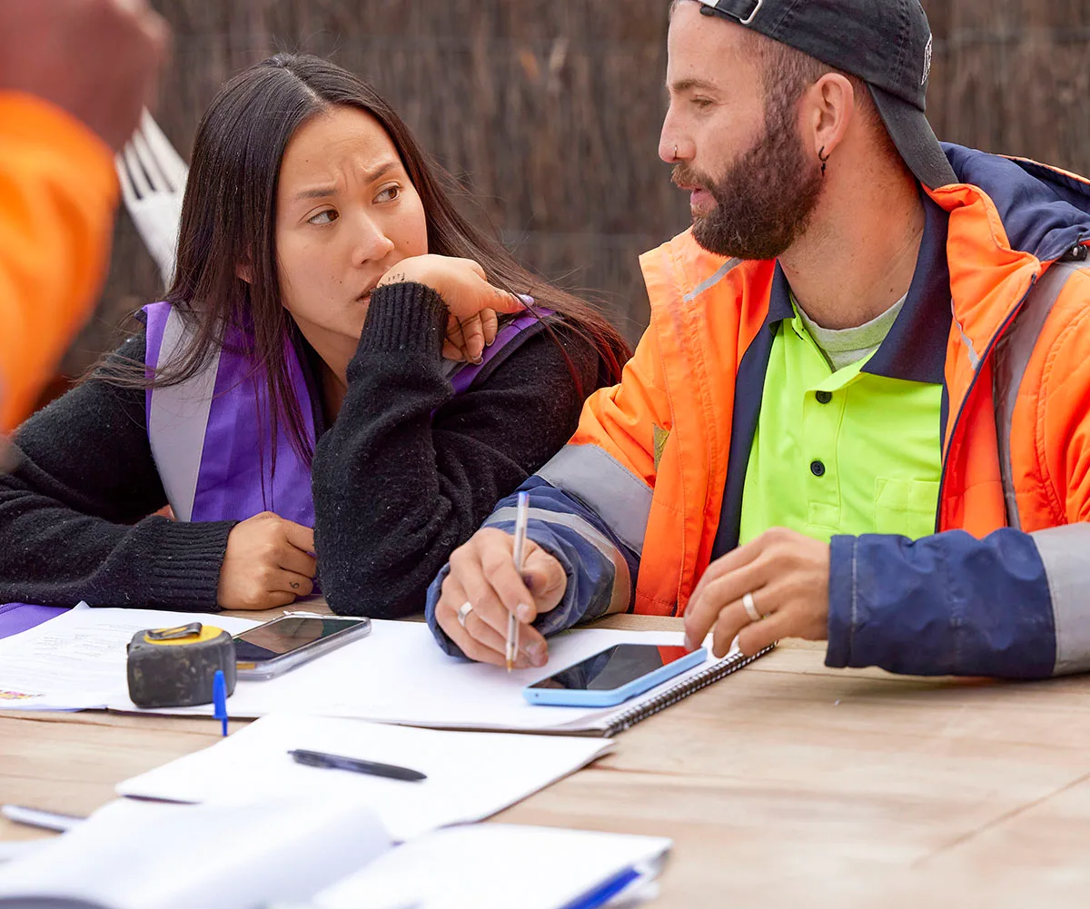 Mimi in purple hi vis and Kristian in orange hi vis sit at a table going over some paperwork looking stressed