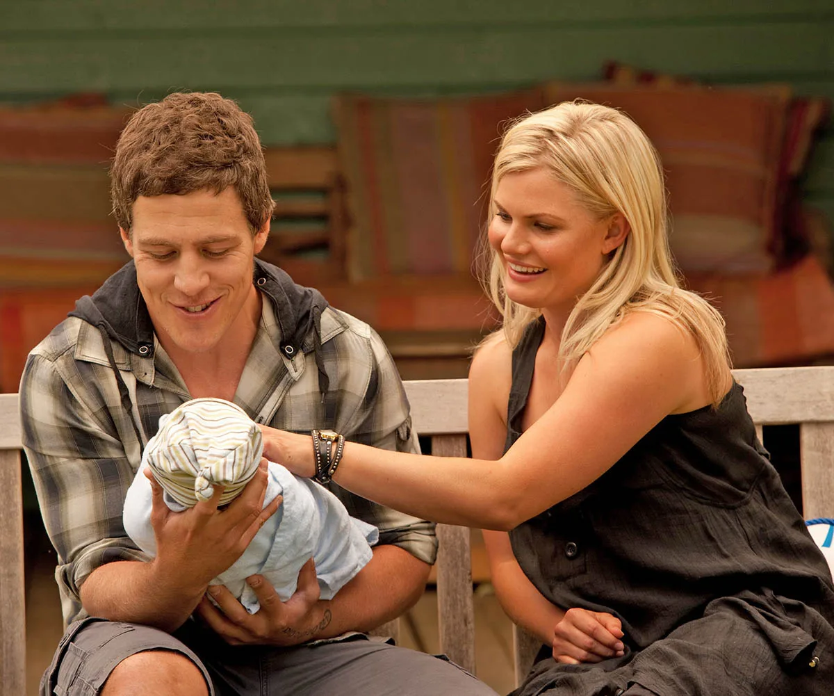 Stephen Peacocke as Brax and Bonnie Sveen as Ricky in a scene from Home And Away. Brax is holding a baby and Ricky is looking affectionately at it