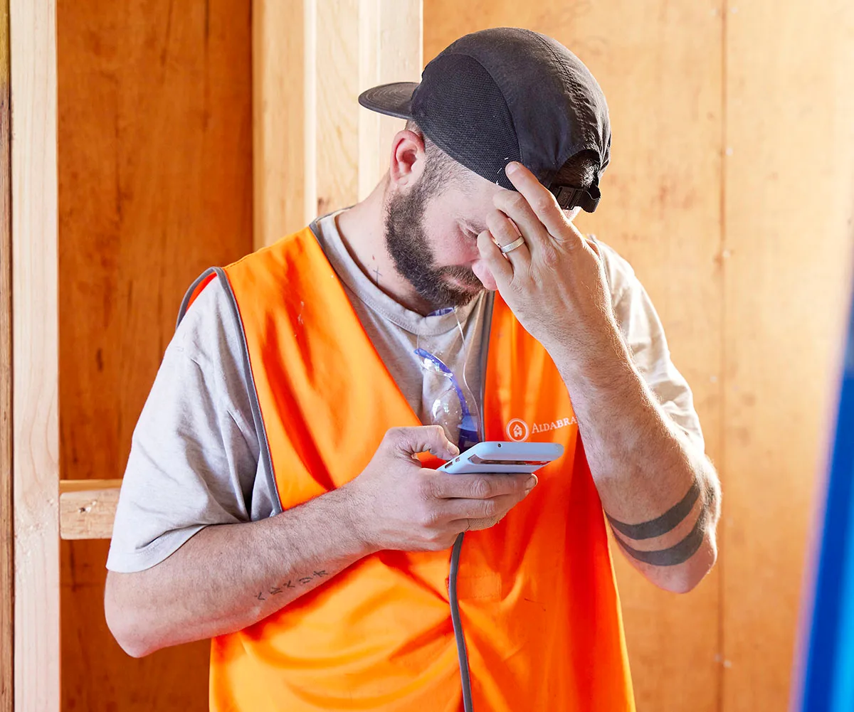 Kristian in orange hi vis and a backwards hat puts a hand to his head as he looks at his phone