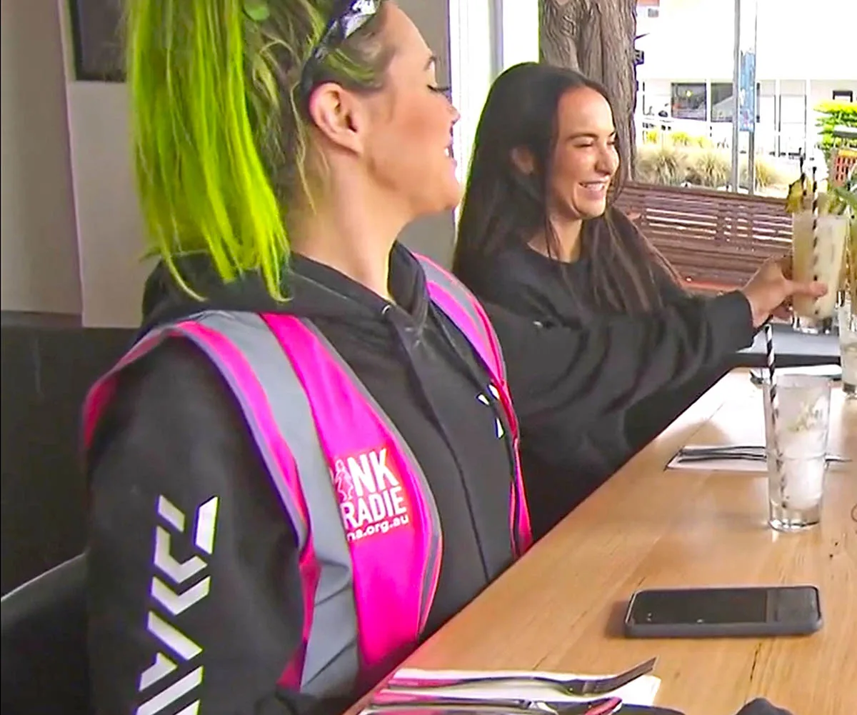 Kylie in pink hi vis and green hair cheersing a pina colada with Courtney with brown hair and black jumper at restaurant table