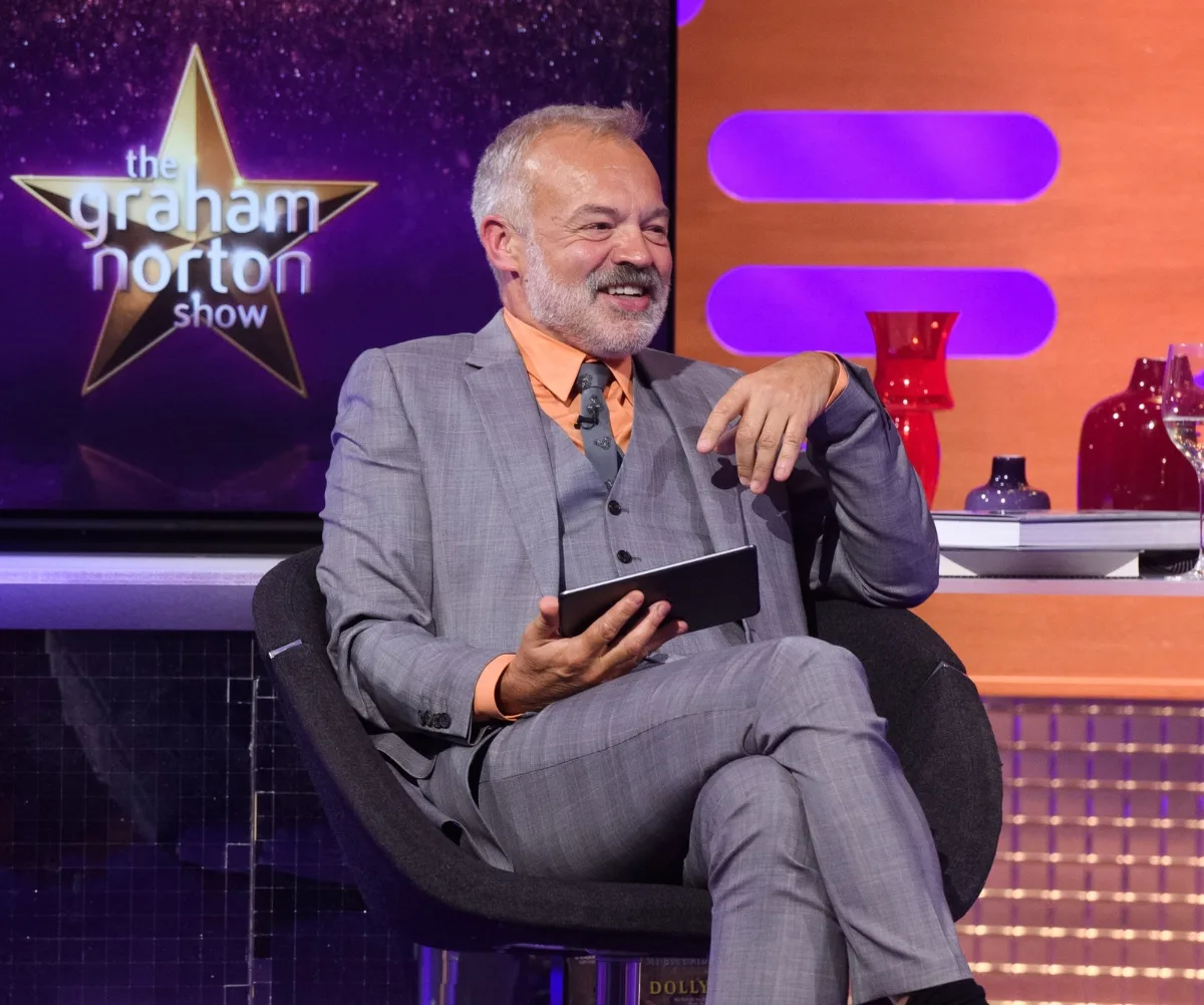 Graham Norton is sitting on a chair laughing while on his chat show