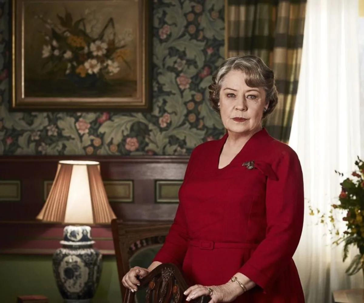 Noni Hazlehurst is wearing a red dress and standing in a formal dining room wearing 1940s costume.