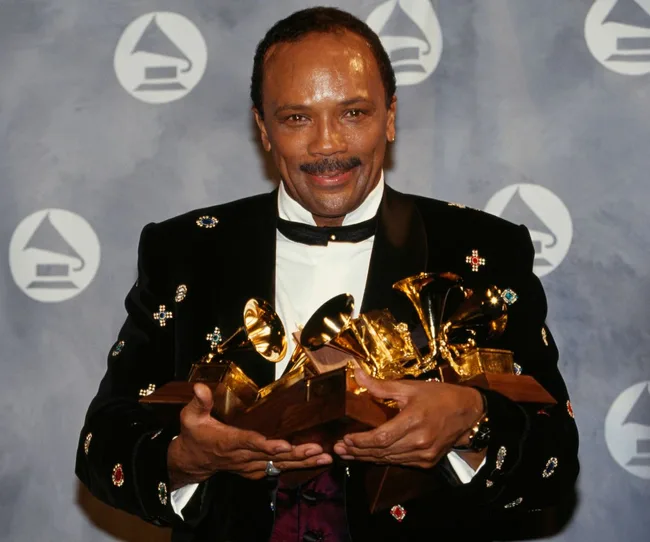 Man holding multiple Grammy awards, wearing a decorated black jacket, smiling at an event with Grammy logo background.