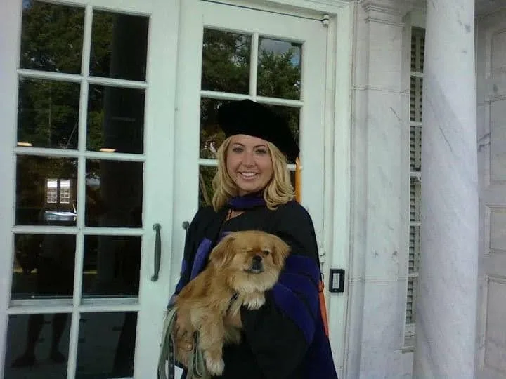 Blonde Lauren Giddings, who was murdered by Stephen McDaniel, in black graduation cape and hat, holding her small, golden dog Butterbean in front of white french doors