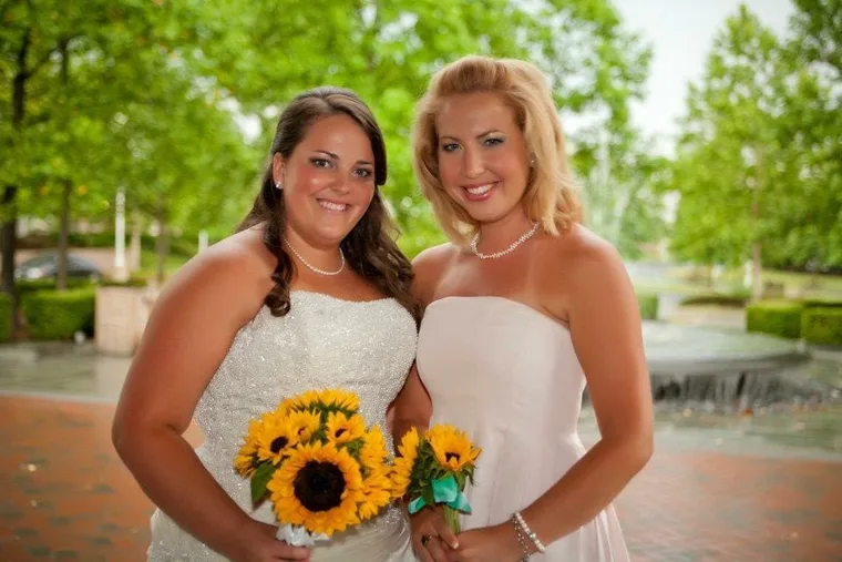 Blonde Lauren Giddings wearing white bridesmaids dress with her brunette sister Kaitlyn Wheeler, wearing white wedding dress holding a bouquet of sunflowers in front of leafy, green background