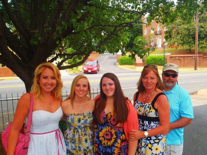 Lauren Giddings in white, sister Sarah in blue and yellow dress, sister Kaitlyn in sunflower patterned dress, mom in blue floral dress and dad in blue polo shirt posing in front of a tree