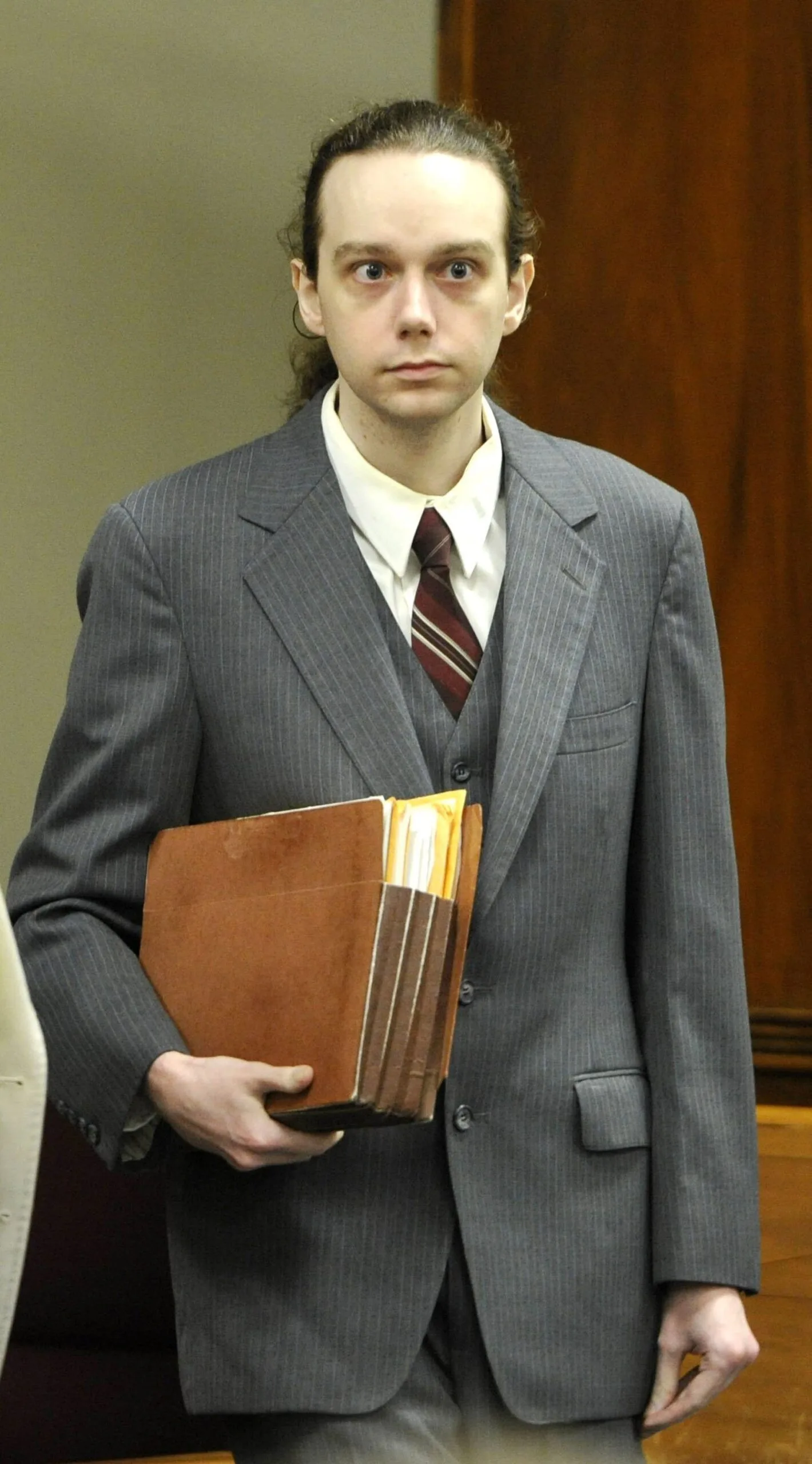 Wide-eyed Stephen McDaniel with long brown hair in ponytail, wearing a grey, pin-stripped suit, white shirt, burgundy tie, carrying a brown filo-folder of documents