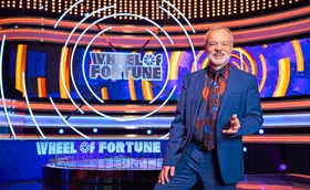 Graham Norton, in a blue suit, stands in front of the Wheel Of Fortune set.