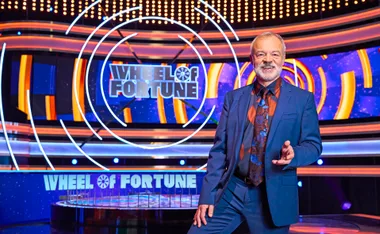 Graham Norton, in a blue suit, stands in front of the Wheel Of Fortune set.