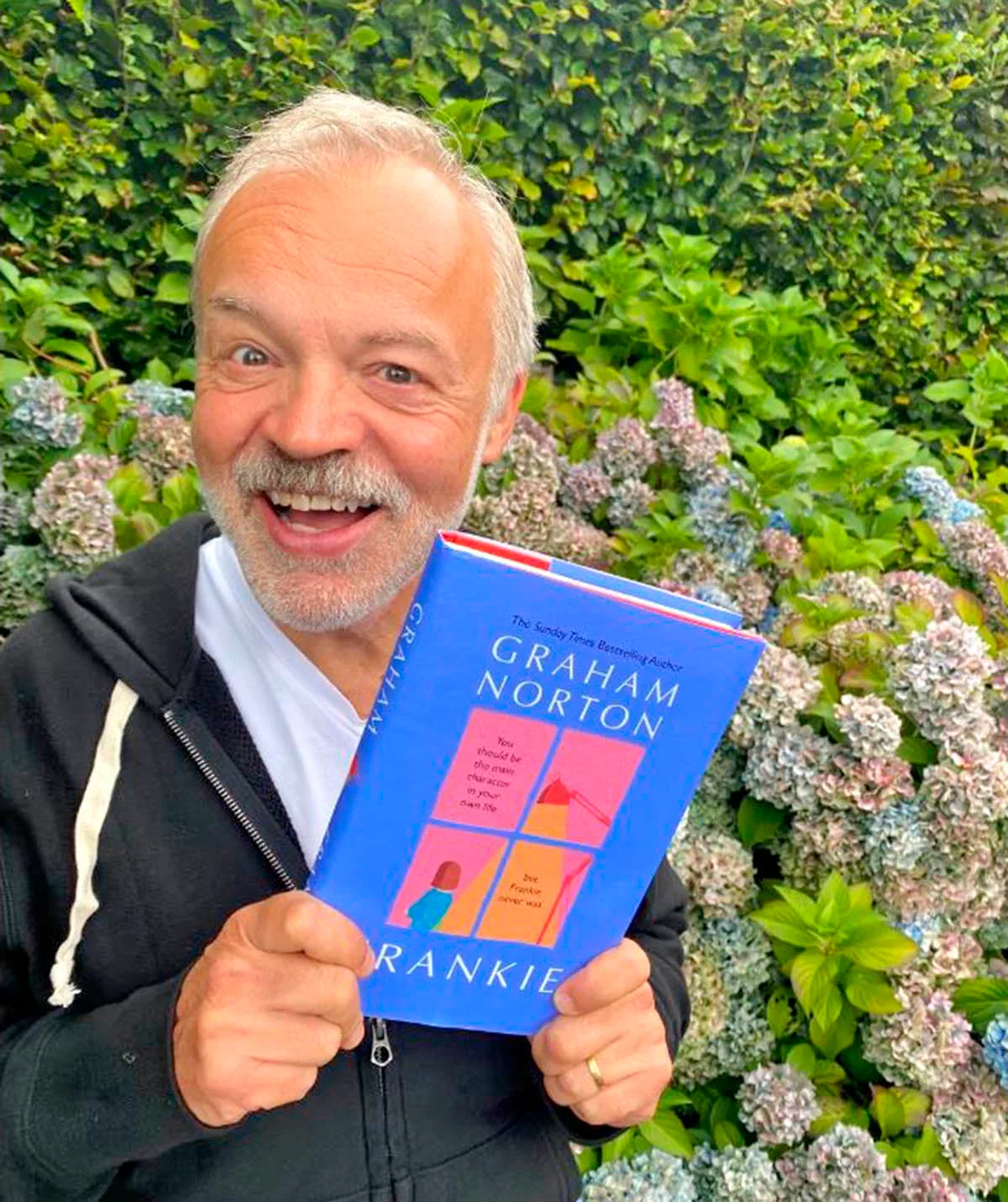 Graham holds a copy of his book, Frankie, while standing in front of a bush.