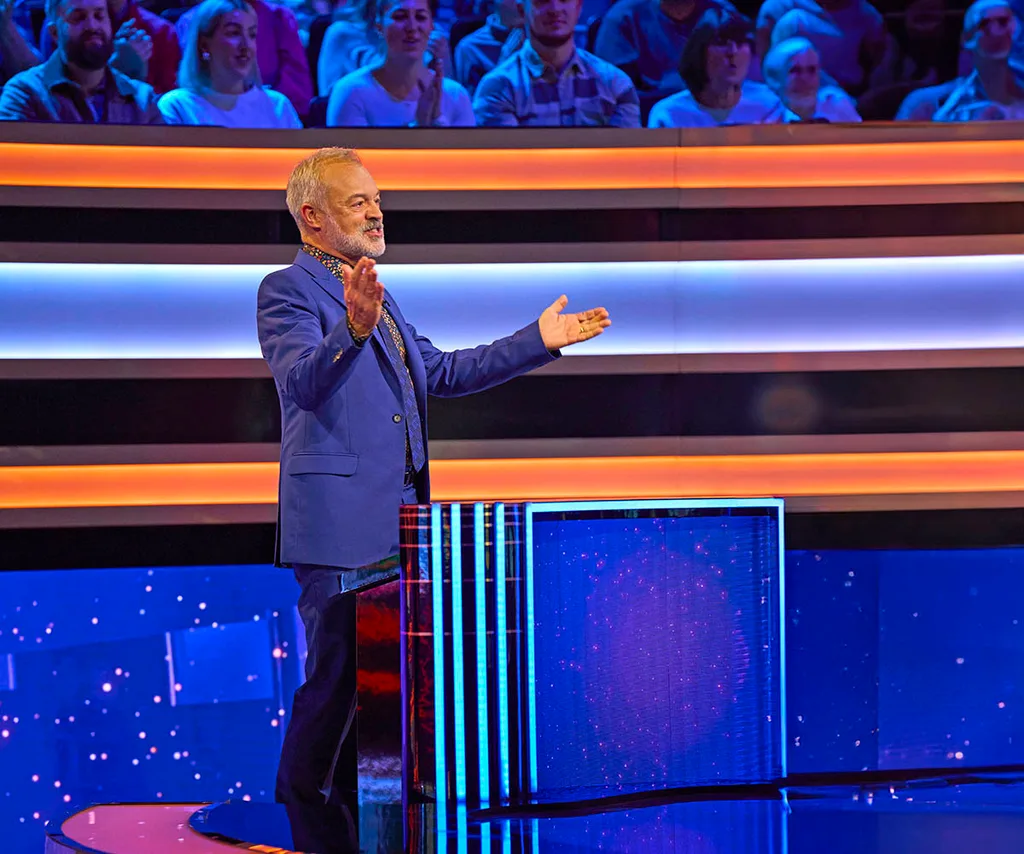 Graham Norton is standing with arms outstretched on the Wheel Of Fortune set.