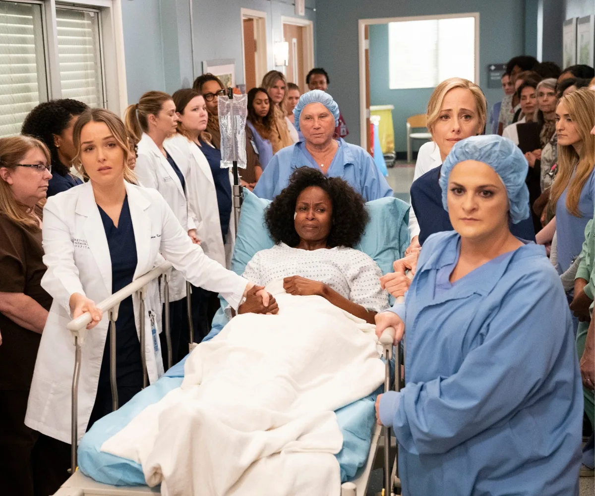 Cast members from Grey's Anatomy are pictured standing around a woman in a hospital bed.