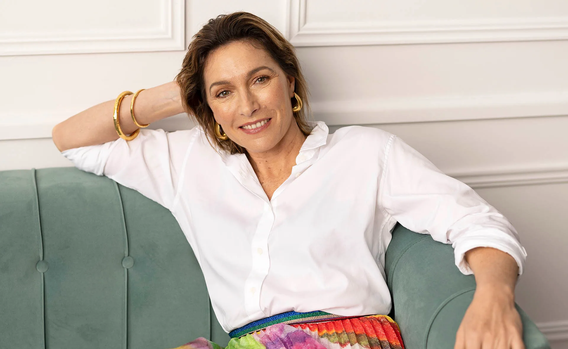 A woman in a white blouse and colorful skirt sits on a green couch, smiling, with a hand resting on her head.