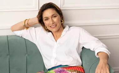 A woman in a white blouse and colorful skirt sits on a green couch, smiling, with a hand resting on her head.