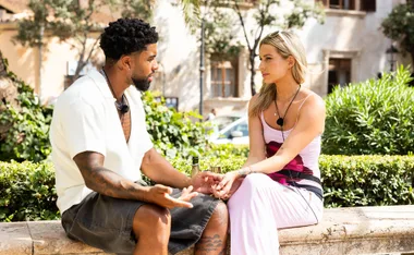 Dylan and Sophie sit facing each other on a ledge in a Spanish square as the talk about their future