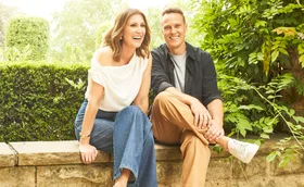 Natalie Barr and Matt Shirvington are sitting next to each other on a low wall and smiling.