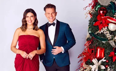 A woman in a red dress and a man in a tuxedo stand smiling beside a decorated Christmas tree.