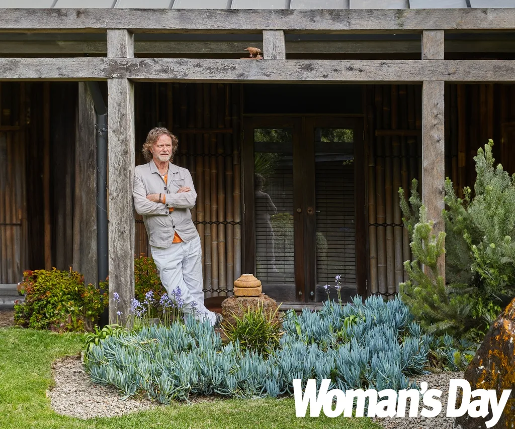 A man stands with his arms crossed leaning against a wooden torii gate in a garden.