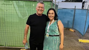 A man in a black shirt and a woman in a green dress smile at the camera, standing outdoors near a fence.