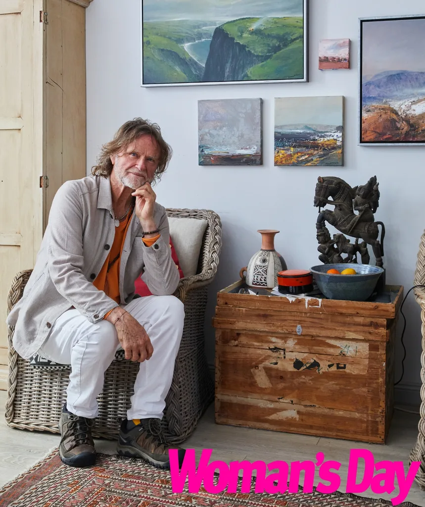 An image of an older man who is sitting down next to a wall covered with paintings.