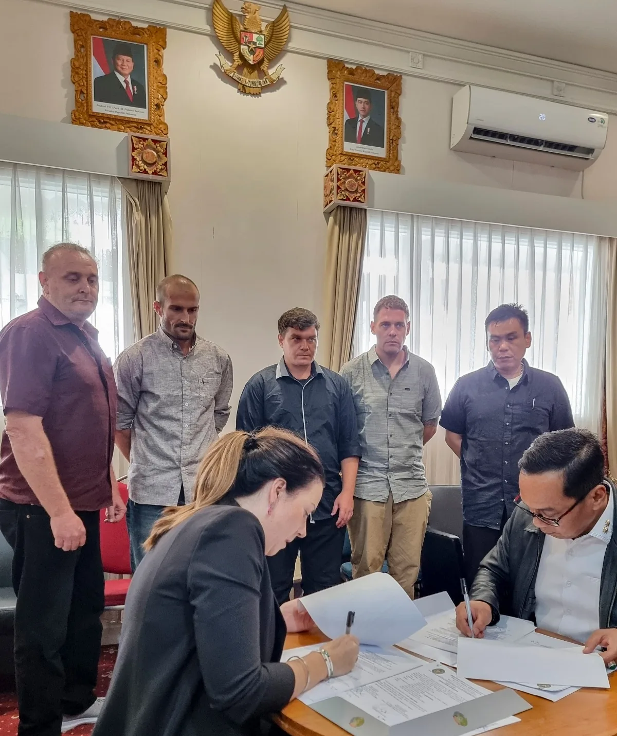 The five members of the Bali Nine stand in a line watching a woman and man sign paperwork.