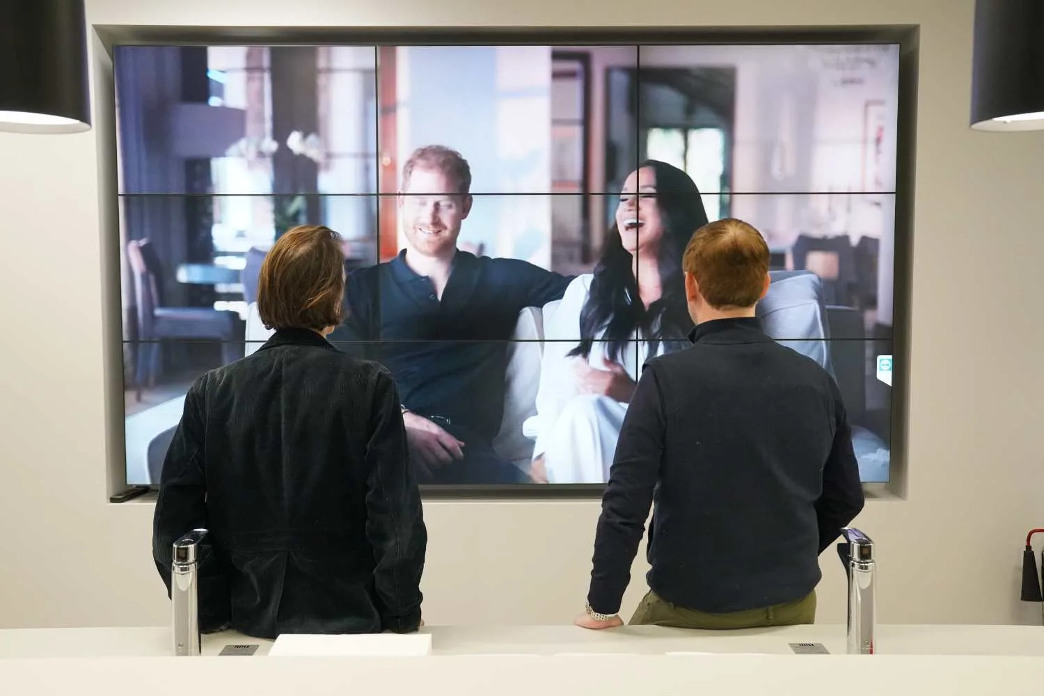 Two people standing in front of screen watching Harry and Meghan 