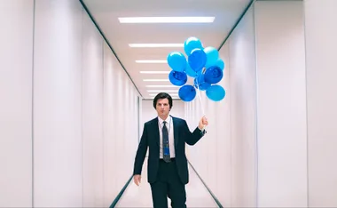 Mark S. holds a bunch of blue balloons in the Lumon Industries white hallway.