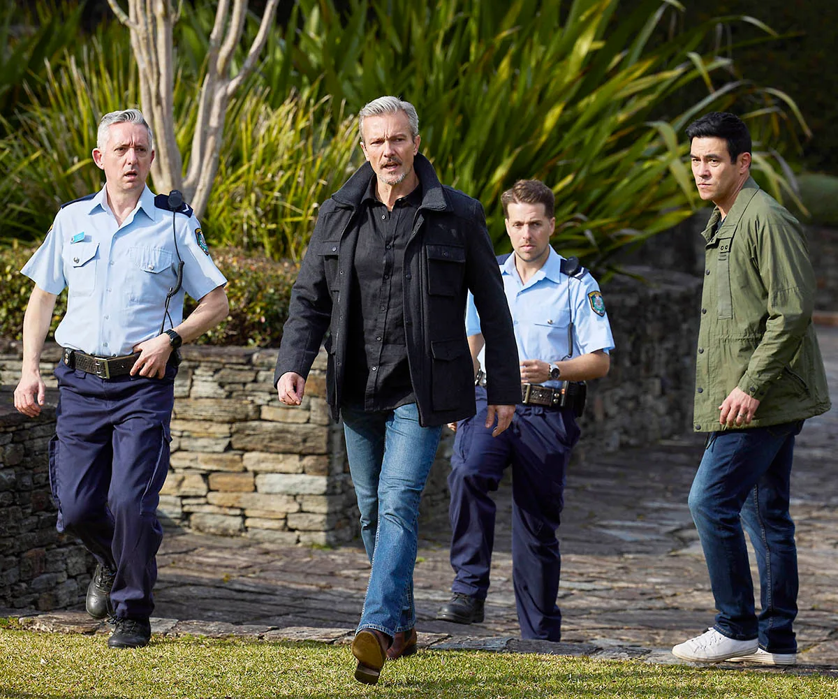 Craig Hall as Detective Townshend leads a team of police across a path of grass as Justin (James Stewart) watches on with a stern look on his face.