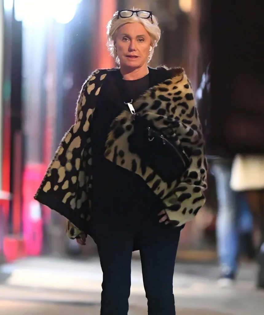 A blonde woman looks at the camera. She is wearing dark clothing and is walking on a city street.