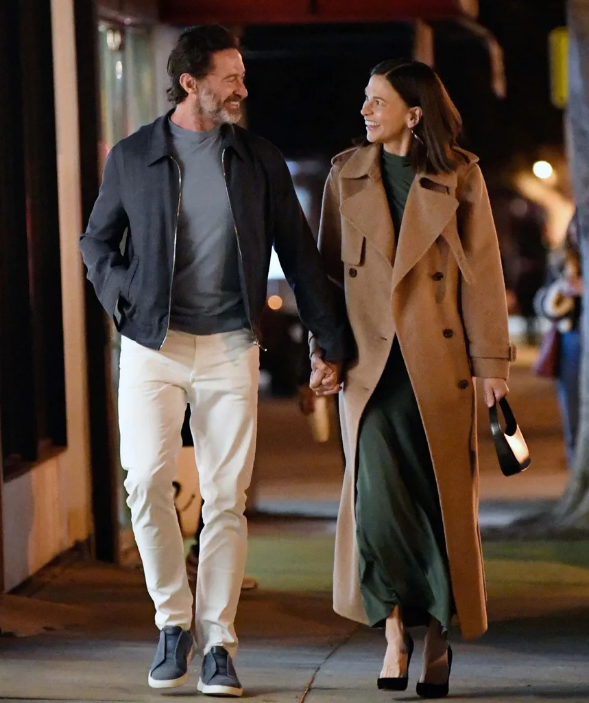 A man and woman walk down the street holding hands and smiling at each other at night time.