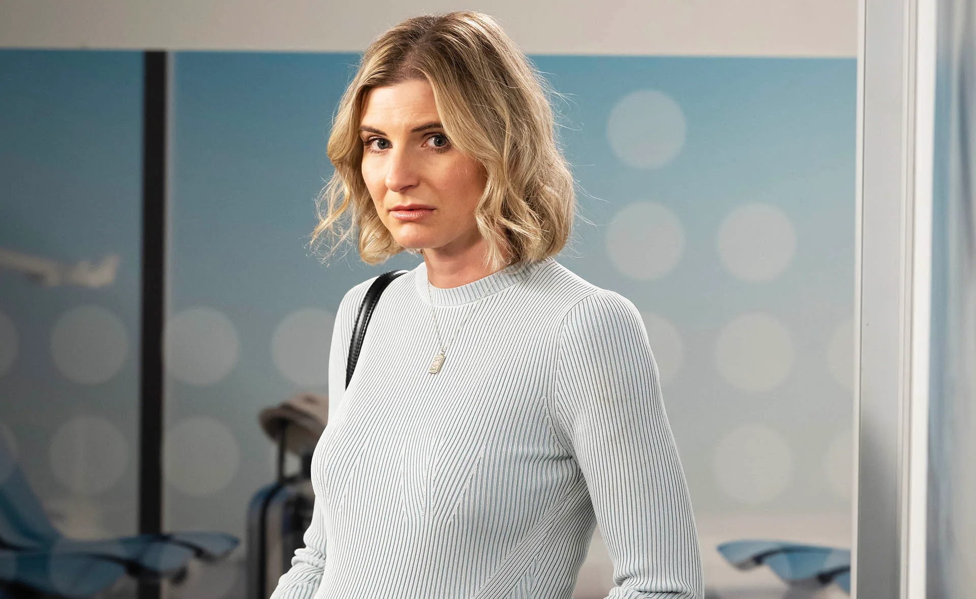 Image of Chelsea in the airport with short blonde hair waved in a tight blue knit long sleeve dress.
