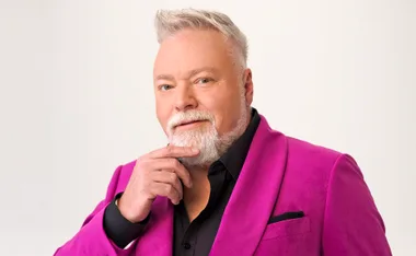 Kyle Sandilands smirks at the camera while grazing his chin with his hand while wearing a black button up shirt and hot pink blazer