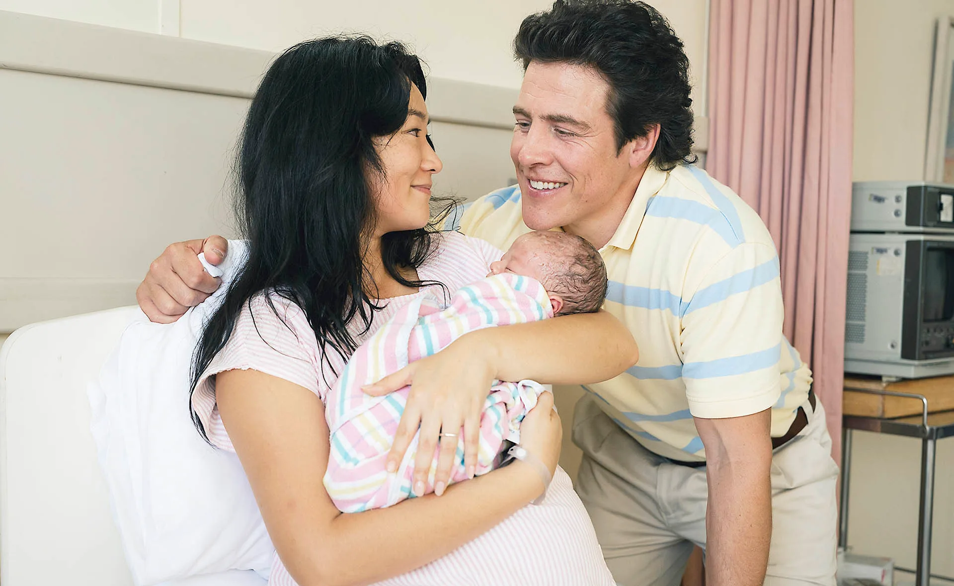 Michelle Lim Davidson, as Noelene, and Stephen Peacocke, as Rob, smile at each other as Noelene holds their newborn baby in a hospital room in The Newsreader