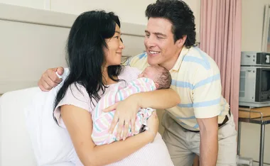 Michelle Lim Davidson, as Noelene, and Stephen Peacocke, as Rob, smile at each other as Noelene holds their newborn baby in a hospital room in The Newsreader