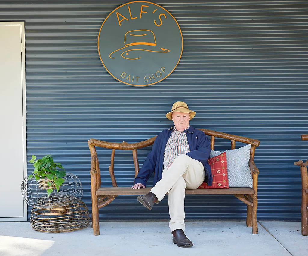 Ray Meagher out the front of a brand new baitshop sitting on a chair.